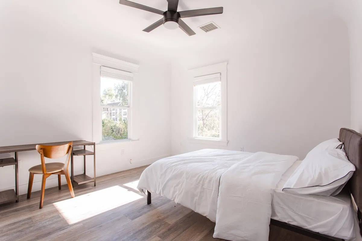 Furnished bedroom with sunlight across the floor, two tall windows with tree views, a queen bed with white bedding, a desk and a wood chair