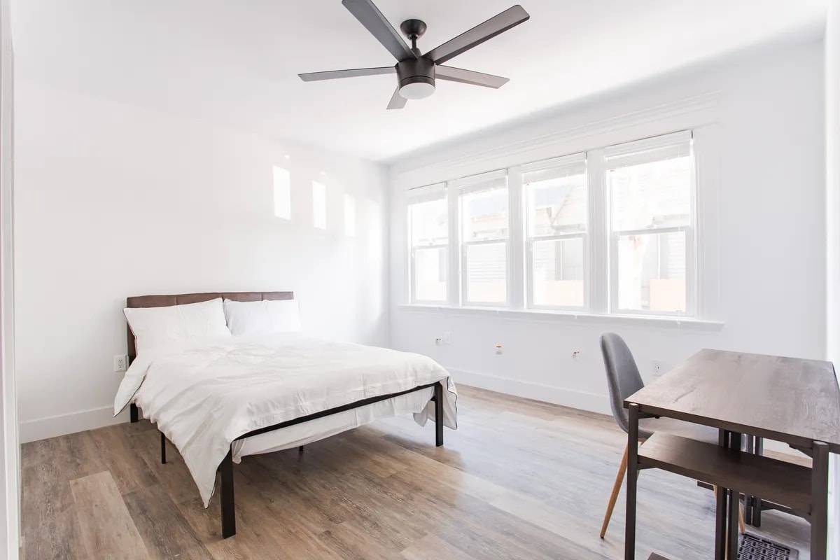 Bedroom with a bank of four windows, a ceiling fan, a bed with white bedding and a desk with a grey chair