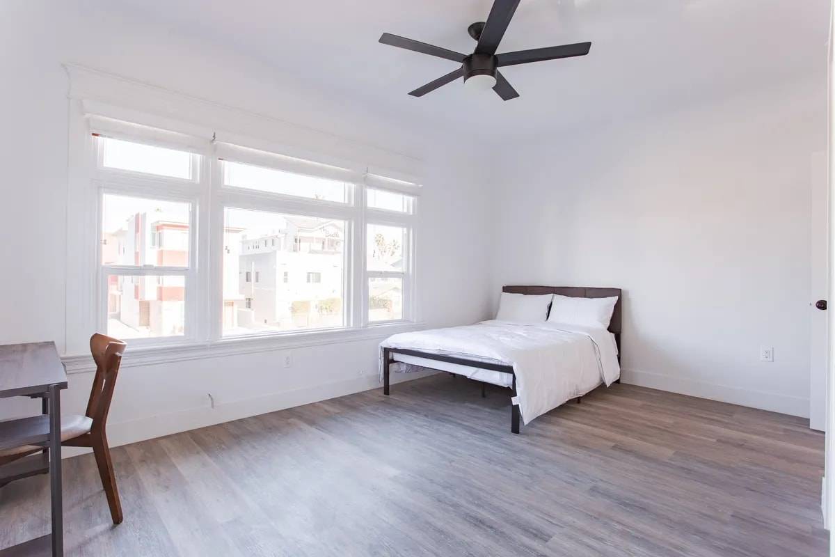 Large bedroom with a wide triple window looking onto the neighborhood, a ceiling fan, a bed and a desk with a wood chair