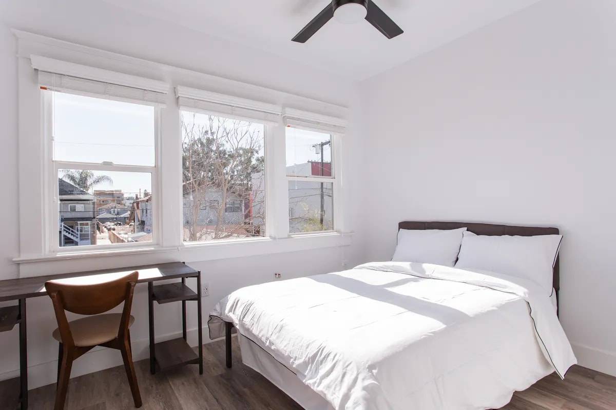 Bedroom with three windows looking out over rooftops and palm trees, a bed with white bedding and a desk with a wood chair
