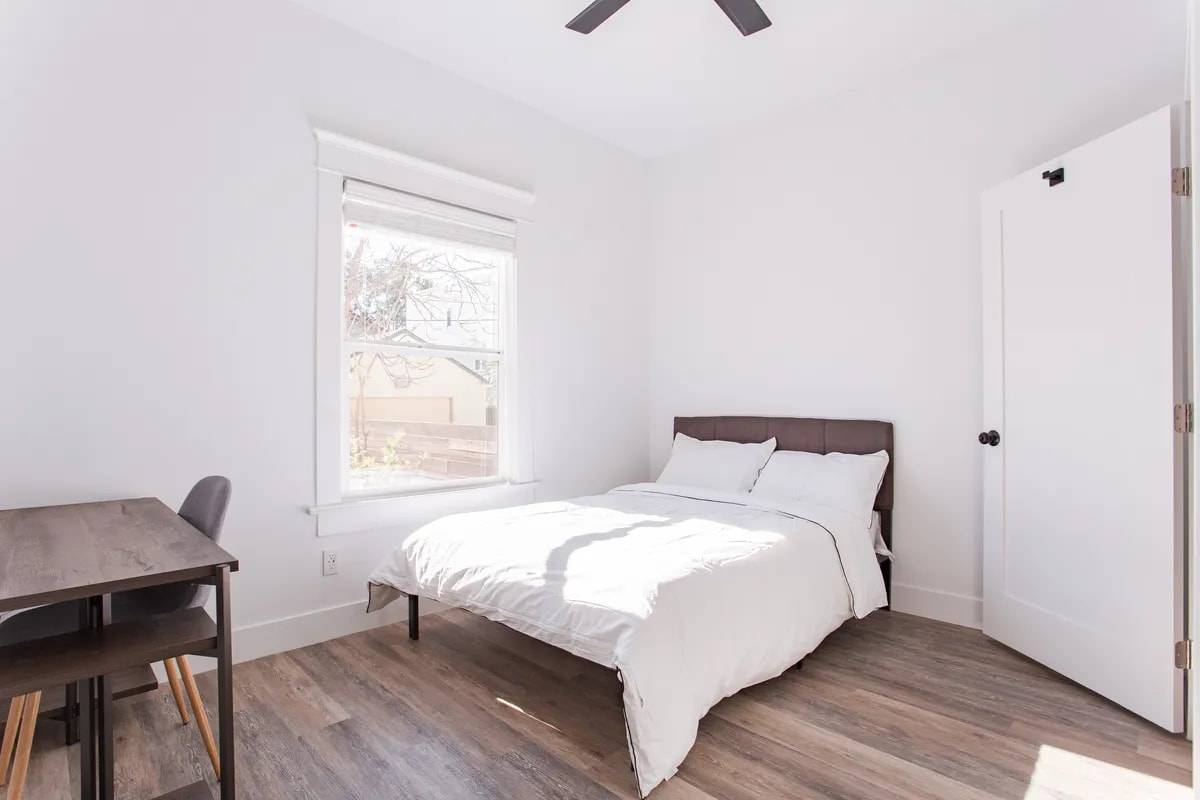 Bedroom with a tall window, a bed with white bedding, a desk and chair, and a white panel door
