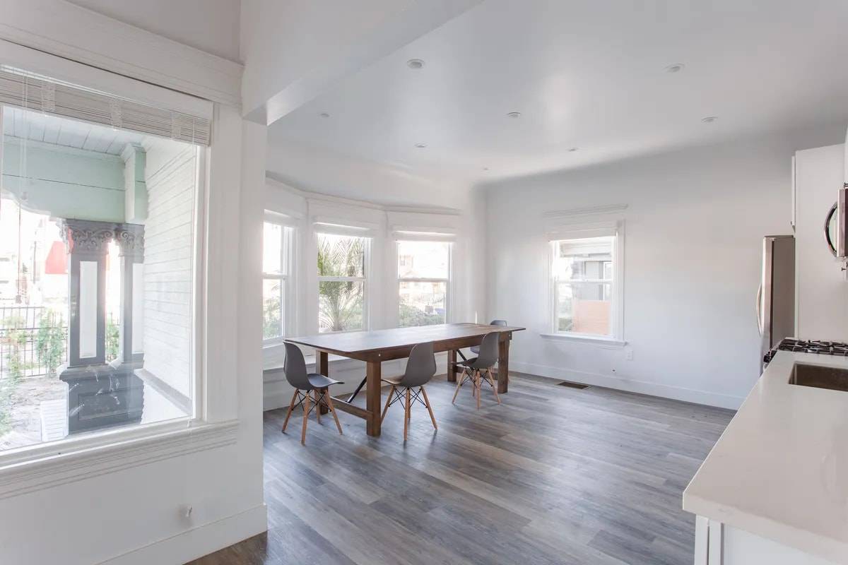 Bright living and dining room with a bay of three windows, a long wood dining table with grey chairs and wood-look plank floors
