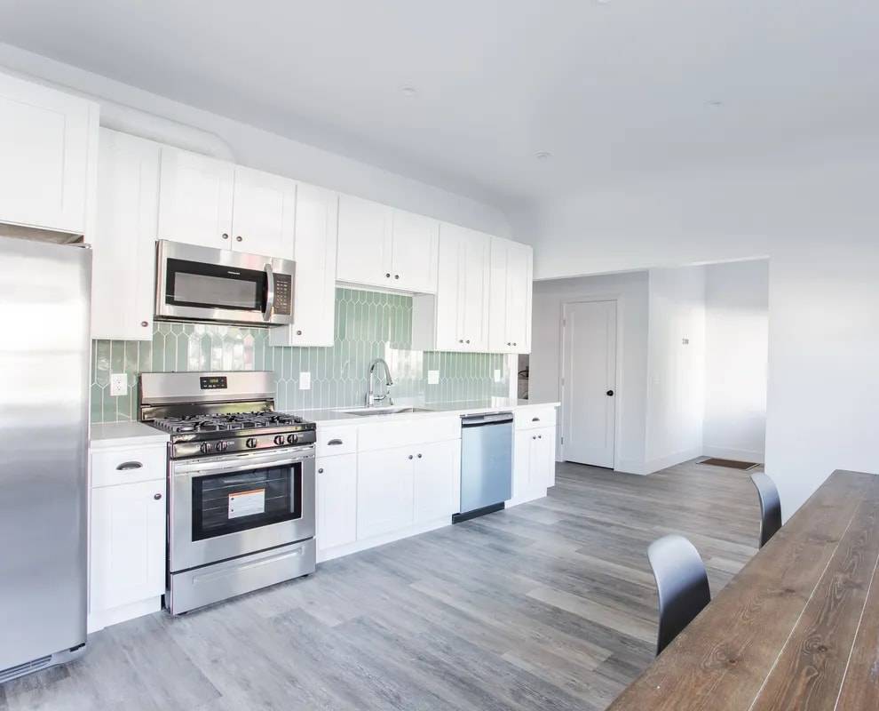 Long galley kitchen with white cabinets, green tile backsplash, stainless refrigerator, range and dishwasher, beside a wood dining table