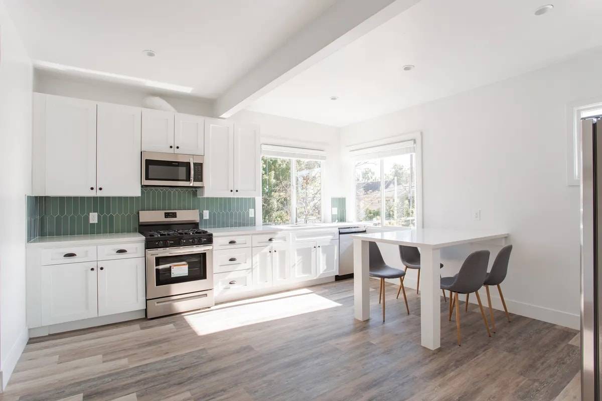 Sunlit kitchen with white shaker cabinets, a sage-green tile backsplash, stainless gas range and microwave, and a white peninsula table with four chairs