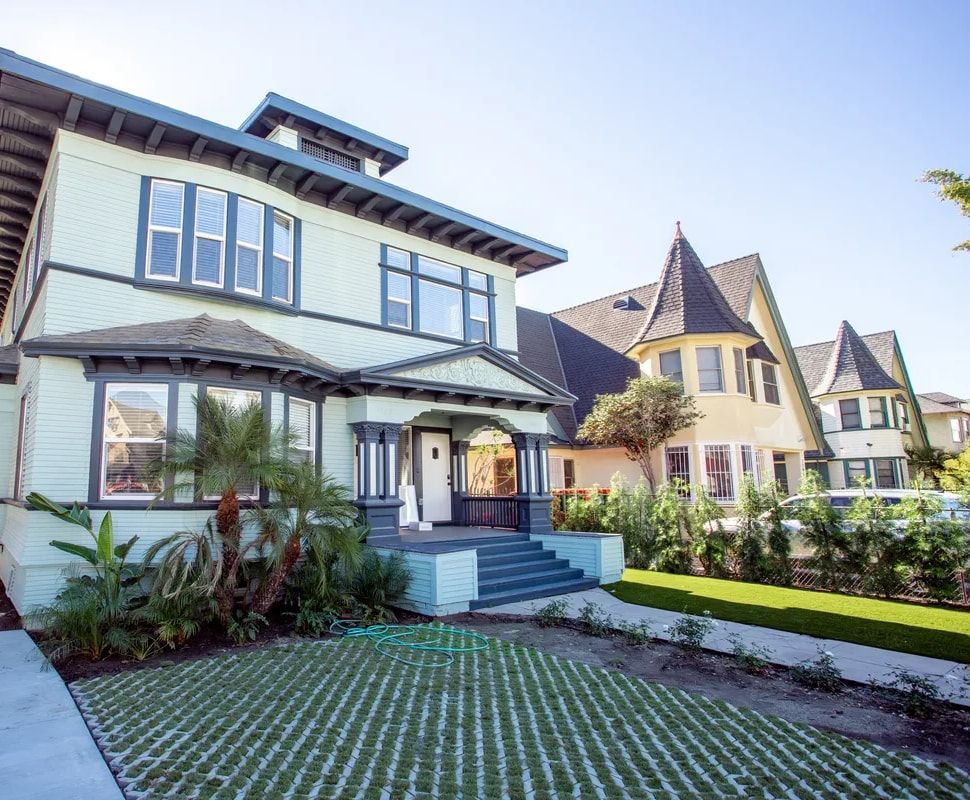 Front of the two-storey 37th Street building: mint-green Craftsman siding with navy trim, a covered front porch with columns, palms and a planted front yard under a clear blue sky